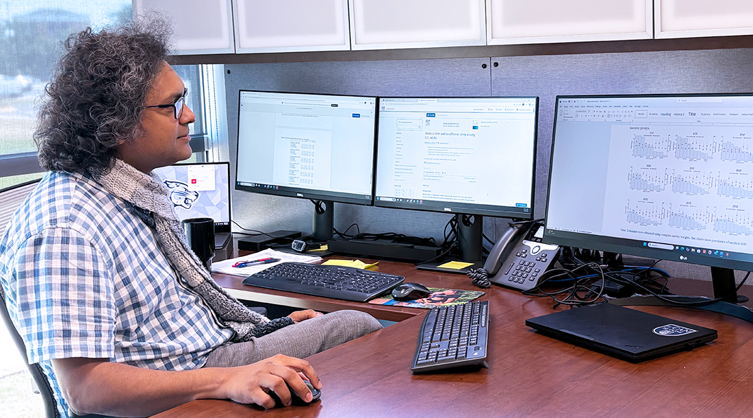 A man wearing a button down short sleeve shirt and a scarf sits in front of a computer and looks at the screen.