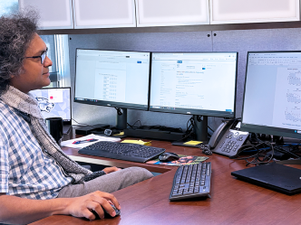A man wearing a button down short sleeve shirt and a scarf sits in front of a computer and looks at the screen.