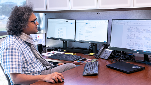 A man wearing a button down short sleeve shirt and a scarf sits in front of a computer and looks at the screen.
