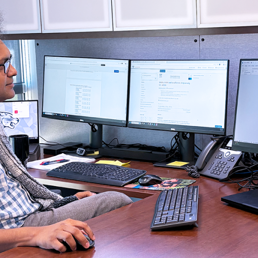A man wearing a button down short sleeve shirt and a scarf sits in front of a computer and looks at the screen.
