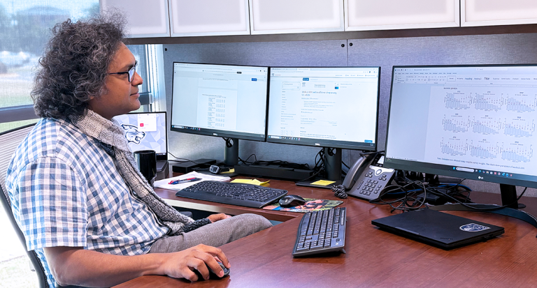 A man wearing a button down short sleeve shirt and a scarf sits in front of a computer and looks at the screen.