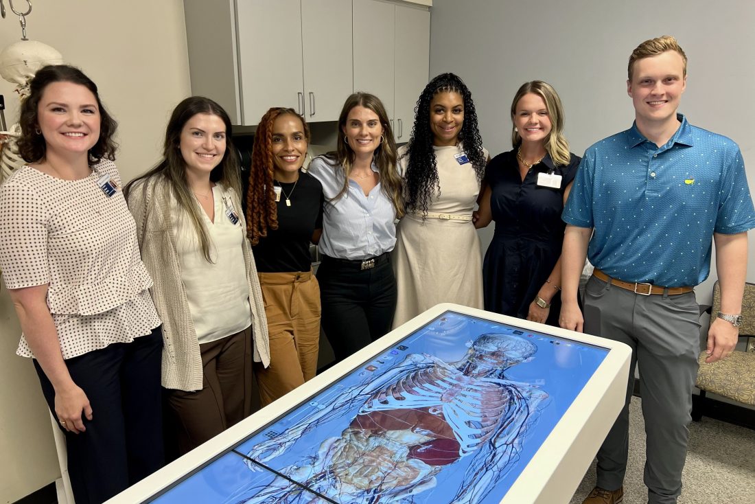 A group of women in business attire pose with a man in a polo. They are all standing behind an advanced anatomy table