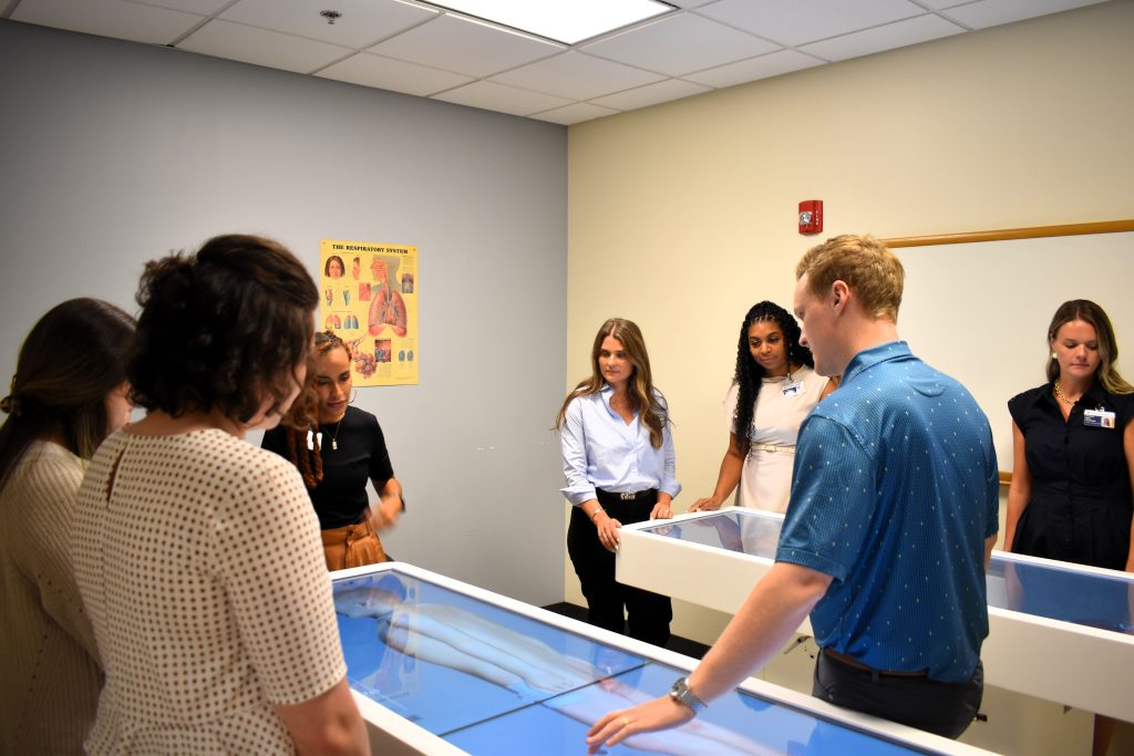 A man leans over a virtual cadaver table, giving a demonstration to a group of students