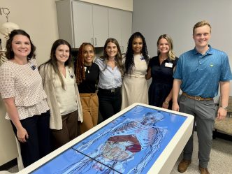 A group of women in business attire pose with a man in a polo. They are all standing behind an advanced anatomy table