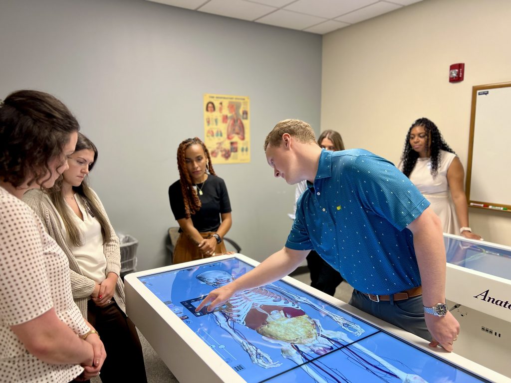 A man leans over a virtual cadaver table, giving a demonstration to a group of students
