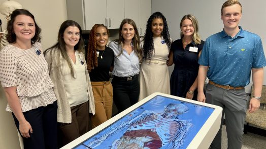 A group of women in business attire pose with a man in a polo. They are all standing behind an advanced anatomy table
