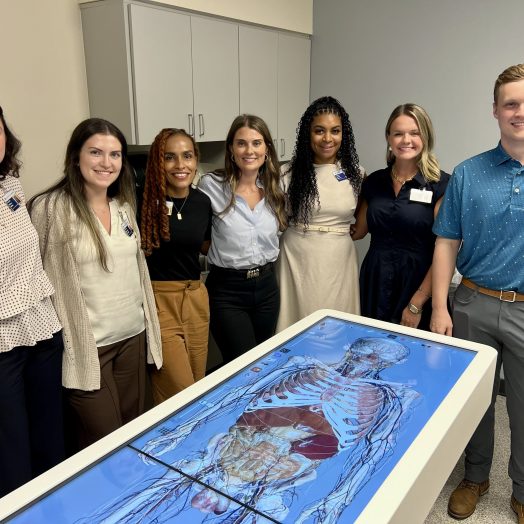 A group of women in business attire pose with a man in a polo. They are all standing behind an advanced anatomy table