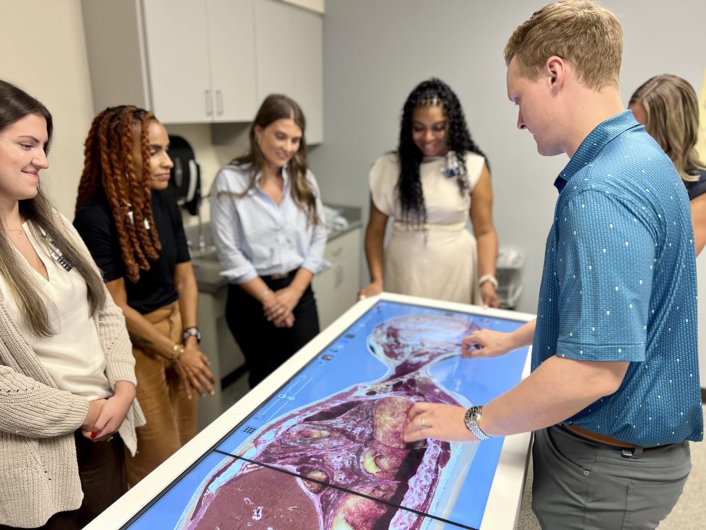 A man leans over a virtual cadaver table, giving a demonstration to a group of students