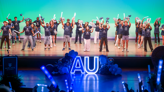 A group of young people on a stage holding pom-poms and glowsticks.