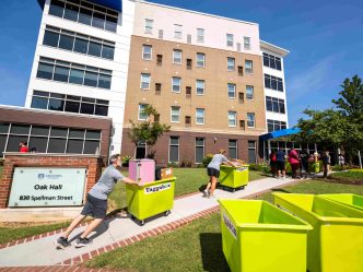 Several men and women push carts filled with students' personal belongings up a small hill, lining up to go into a residence hall.