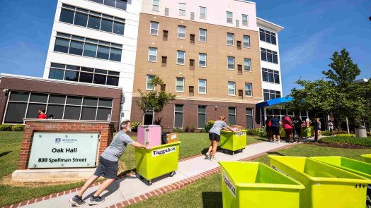 Several men and women push carts filled with students' personal belongings up a small hill, lining up to go into a residence hall.