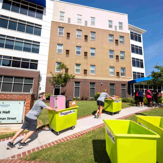Several men and women push carts filled with students' personal belongings up a small hill, lining up to go into a residence hall.