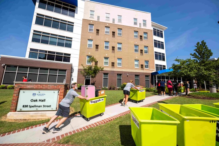 Several men and women push carts filled with students' personal belongings up a small hill, lining up to go into a residence hall.
