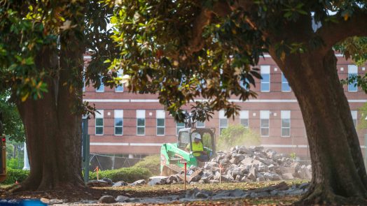 Surrounded by large trees, a mini excavator shovels broken cement off the grass.
