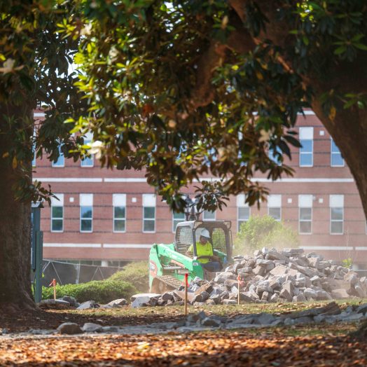 Surrounded by large trees, a mini excavator shovels broken cement off the grass.