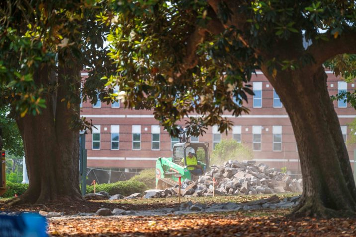 Surrounded by large trees, a mini excavator shovels broken cement off the grass.
