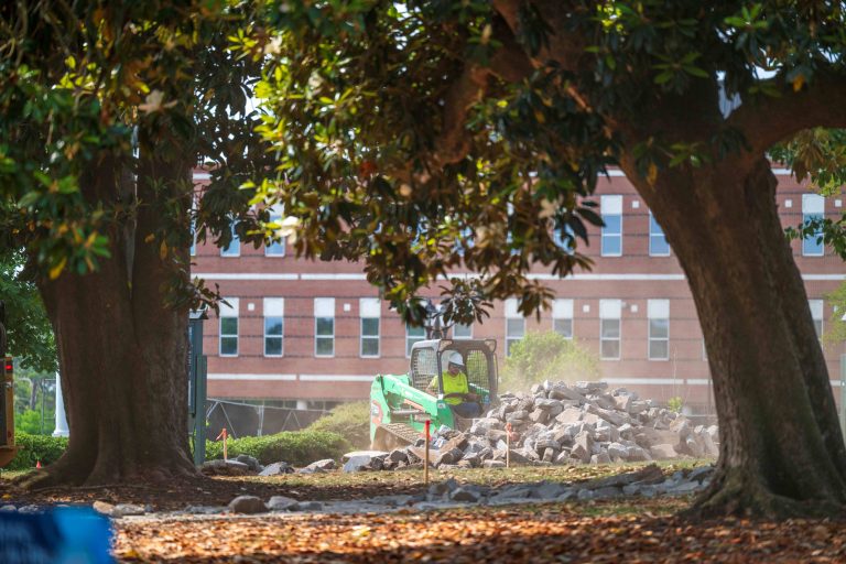 Surrounded by large trees, a mini excavator shovels broken cement off the grass.