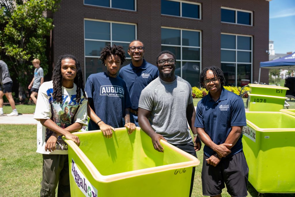While standing outdoors, several men smile near a moving cart. 