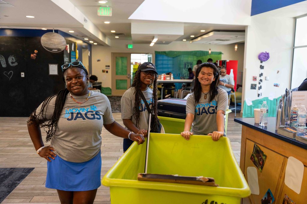 While standing indoors, several women smile near a moving cart. 