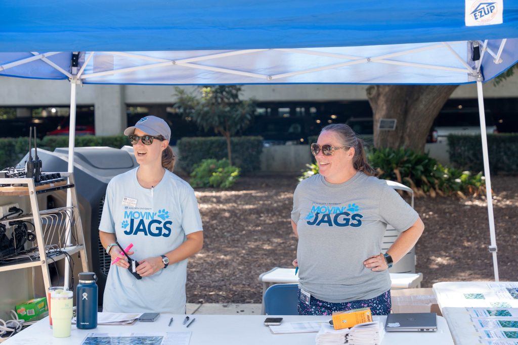 Two women standing under an outdoor tent speak with a person off to the side. 