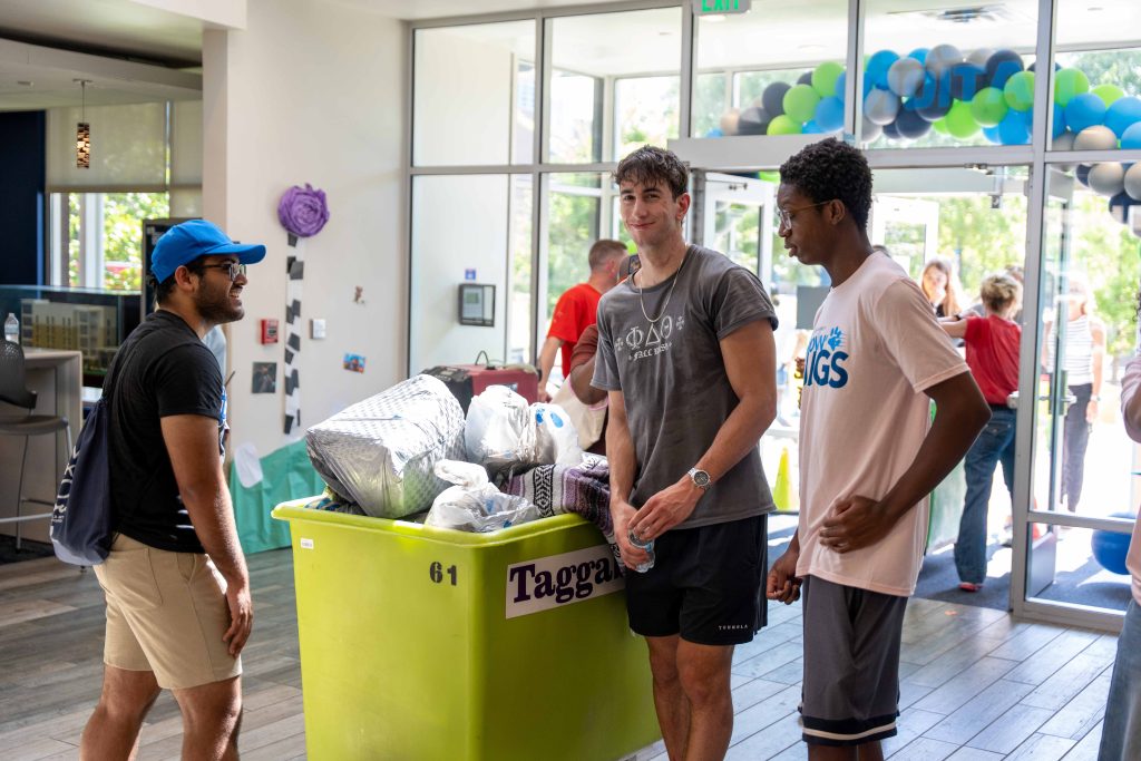 While standing indoors, three college-aged men stand talking near a move-in cart.