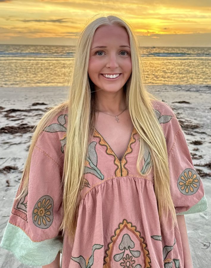 Young woman stands on beach in front of sunset and waves, smiling.