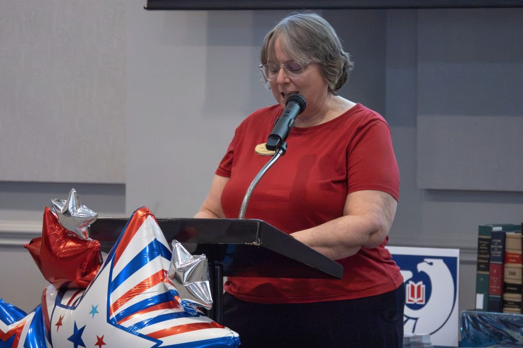 A woman in a red shirt speaks at a podium adorned with balloons. In the background are government books.