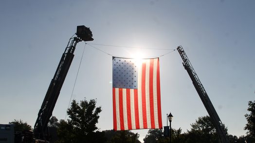 A giant United States flag is hung on two firefighter ladders high above the ground with the sun shining through