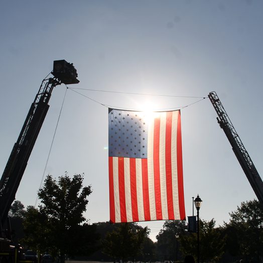 A giant United States flag is hung on two firefighter ladders high above the ground with the sun shining through
