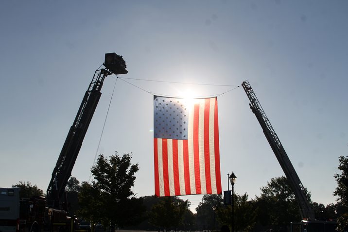 A giant United States flag is hung on two firefighter ladders high above the ground with the sun shining through