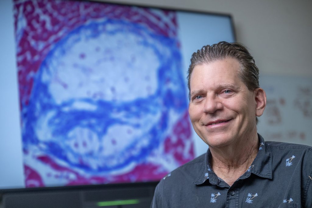 A man in a gray shirt smiles in front of an image of a cell