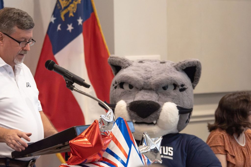 A man speaks at a microphone with the flag of Georgia and the Augusta University mascot in the background.