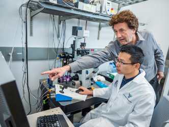 Two male scientists work at a computer station in a lab. One, the senior scientist, leans over the shoulder of the younger scientist to point to figures on the screen.