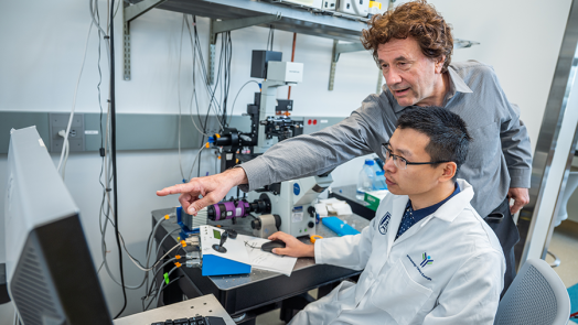 Two male scientists work at a computer station in a lab. One, the senior scientist, leans over the shoulder of the younger scientist to point to figures on the screen.