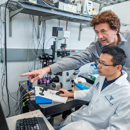 Two male scientists work at a computer station in a lab. One, the senior scientist, leans over the shoulder of the younger scientist to point to figures on the screen.