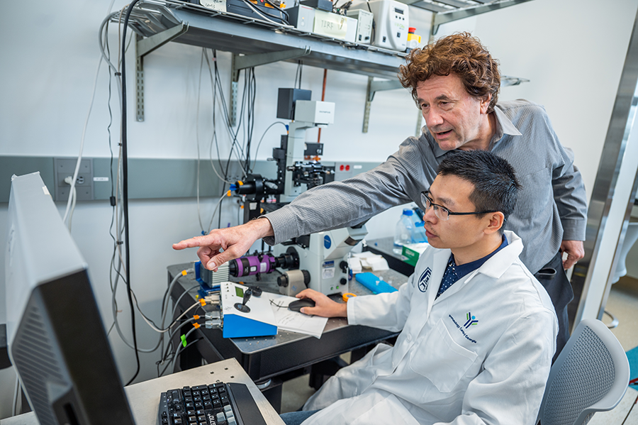Two male scientists work at a computer station in a lab. One, the senior scientist, leans over the shoulder of the younger scientist to point to figures on the screen.