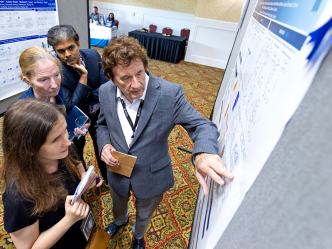 A male scientist discusses points from a scientific poster with a female junior scientist while two other scientists, a man and a woman, look on.