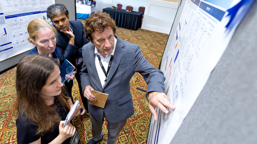 A male scientist discusses points from a scientific poster with a female junior scientist while two other scientists, a man and a woman, look on.