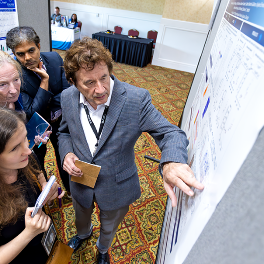A male scientist discusses points from a scientific poster with a female junior scientist while two other scientists, a man and a woman, look on.