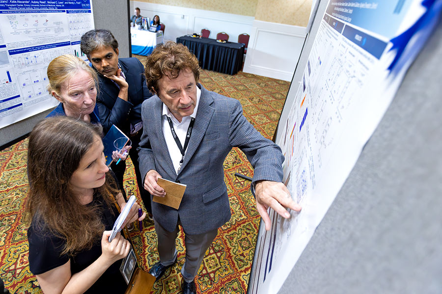 A male scientist discusses points from a scientific poster with a female junior scientist while two other scientists, a man and a woman, look on.