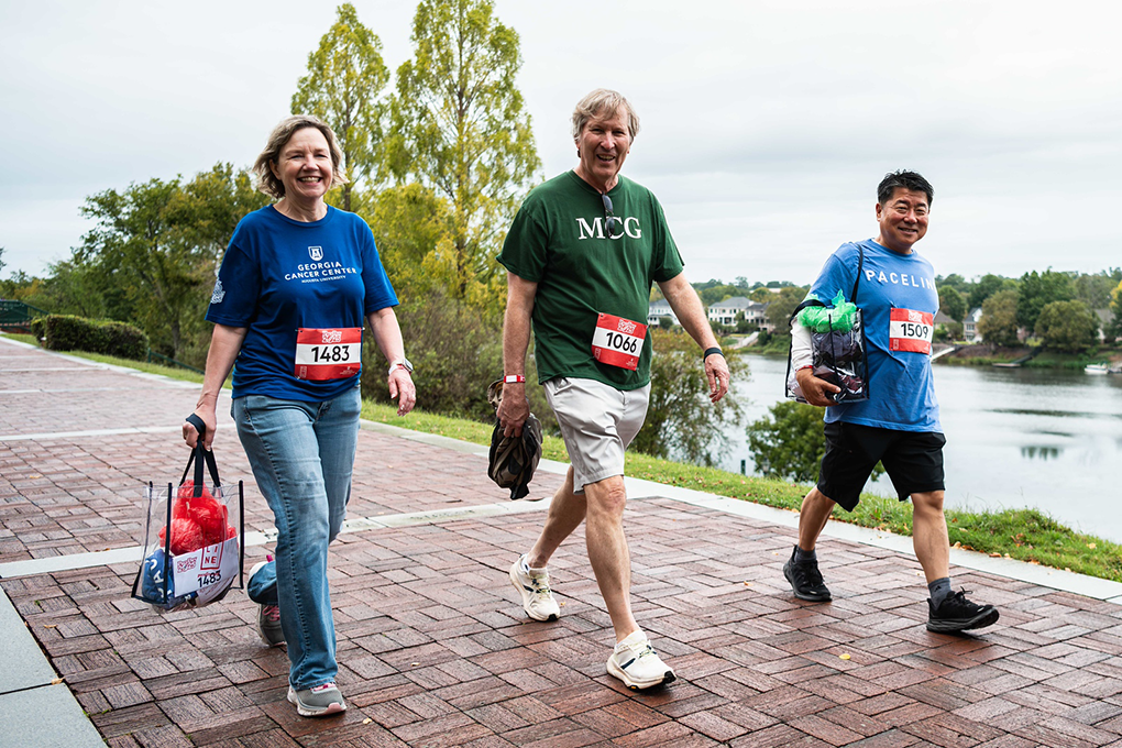 Two men and a woman walk on a brick sidewalk next to a body of water and smile.