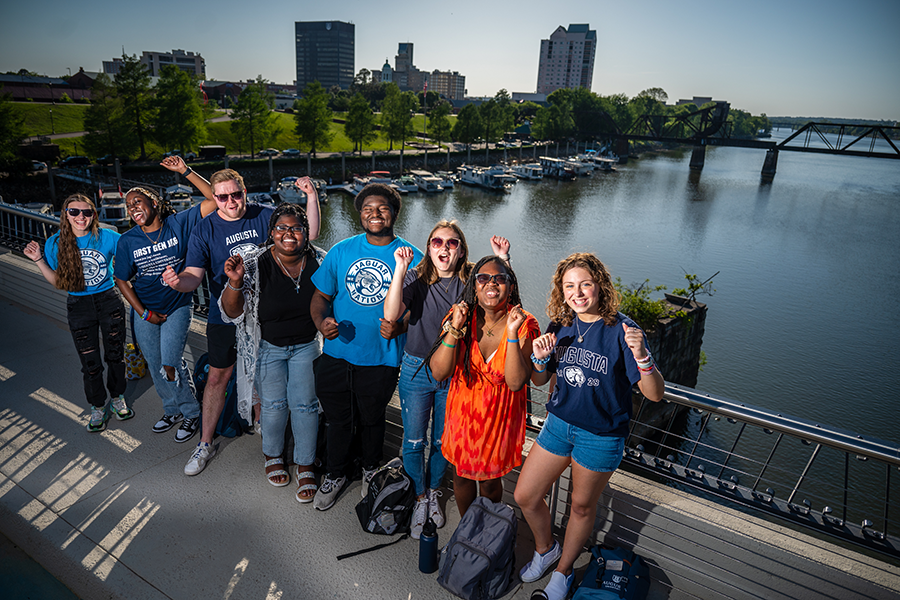 Group of students with their backs against the railing of a bridge face the camera and cheer. 