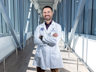 A male scientist in a laboratory coat stands in a long, modern bridge connecting two scientific labs.