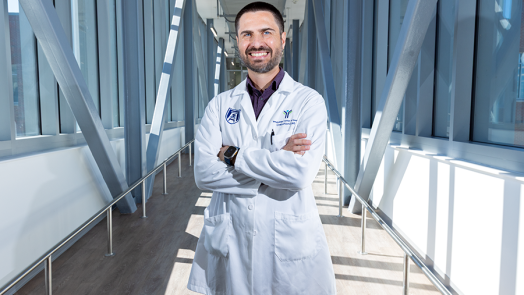 A male scientist in a laboratory coat stands in a long, modern bridge connecting two scientific labs.