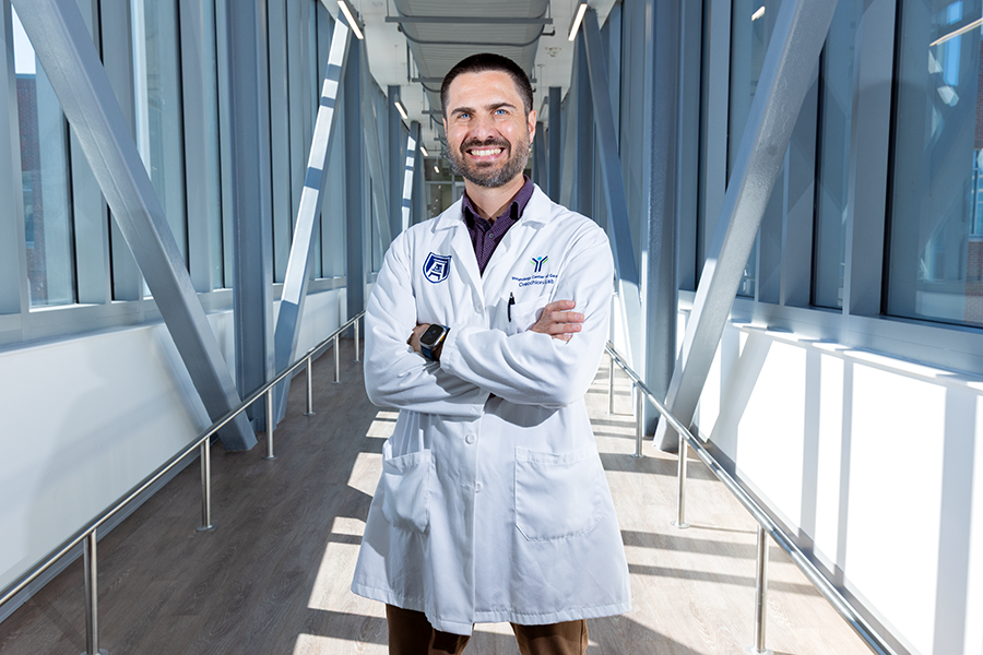 A male scientist in a laboratory coat stands in a long, modern bridge connecting two scientific labs.