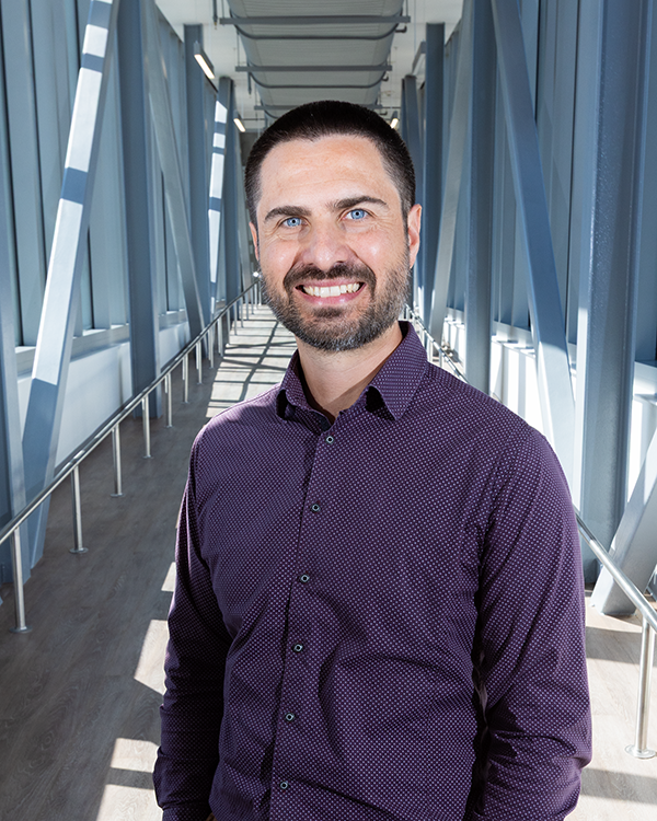 A male scientist stands in a long, modern bridge connecting two scientific labs.