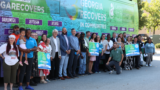 A large group of people stand in front of an addiction recovery bus and smile.