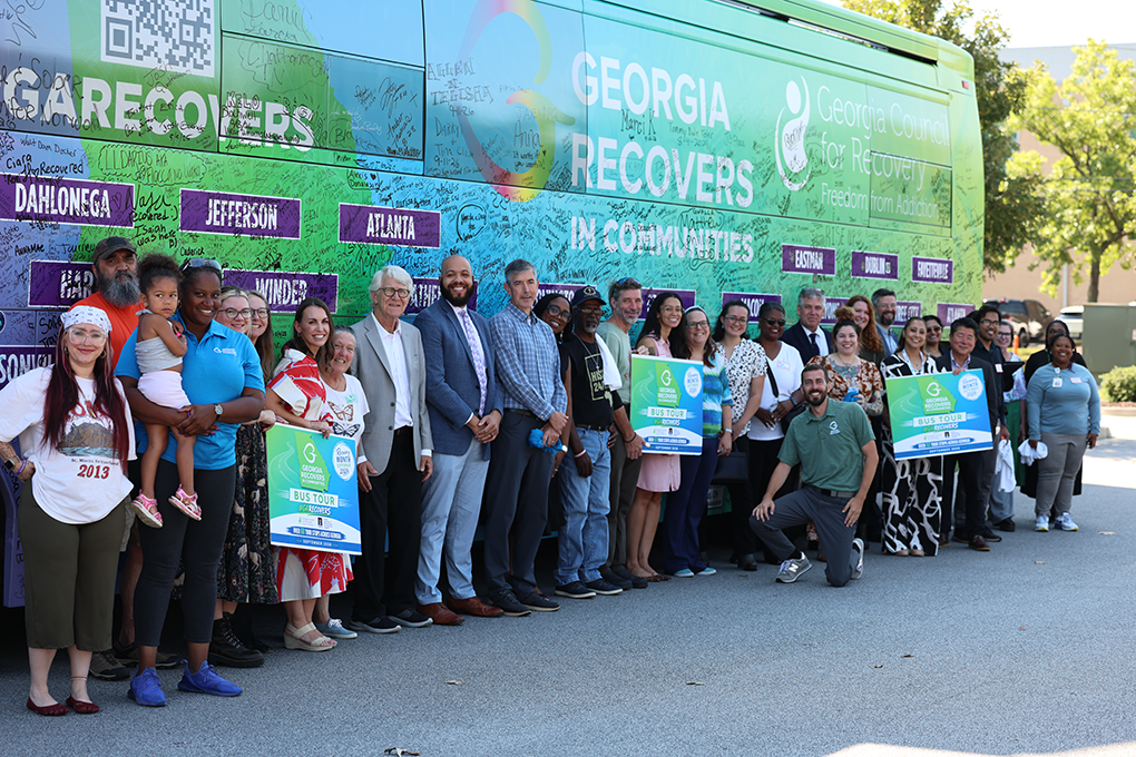 A large group of people stand in front of an addiction recovery bus and smile.