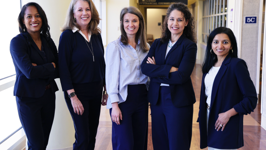 Five professionally dressed woman stand beside each other and smile.