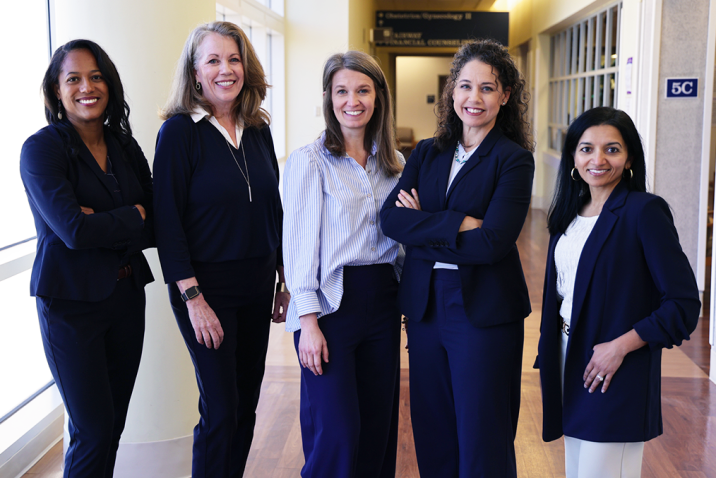 Five professionally dressed woman stand beside each other and smile.